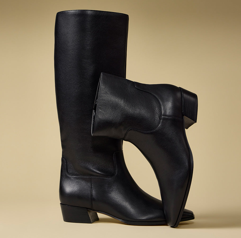 A pair of black leather knee-high boots with a unique asymmetrical design, featuring one boot standing upright and the other tilted in front to show the side profile and heel detail, set against a solid beige background.