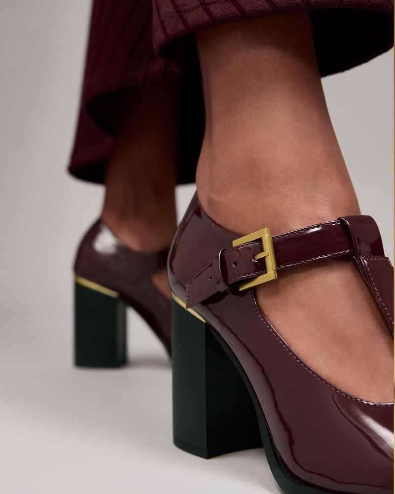 Close-up of a person wearing the Vince Camuto Micha Pump—shiny burgundy heeled Mary Jane shoes with gold buckles—paired with cropped burgundy pants. The focus is on the shoes and lower legs, emphasizing the vintage charm and feminine style of this classic T-strap pump.