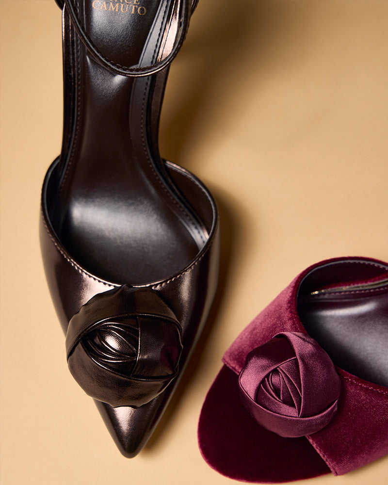 A top view of two Vince Camuto Mirezzo Pumps on a tan surface: one metallic black stiletto with a leather rose and pointed toe, and one burgundy with a fabric rose and rounded toe.