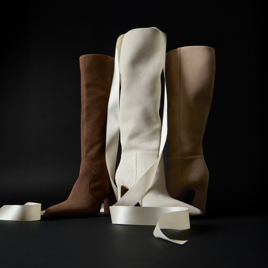 3 neutral-toned suede Knee High Boots shot in a dimly-lit studio with white holiday-ribbon overlayed