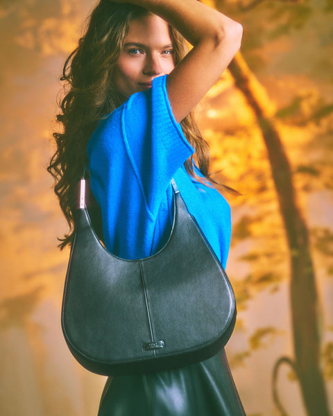 A woman with long hair in a blue top lifts her arm, showcasing the Vince Camuto Noli Hobo Bag— a spacious black purse—over her shoulder. The background features warm, blurred tones and tree silhouettes.