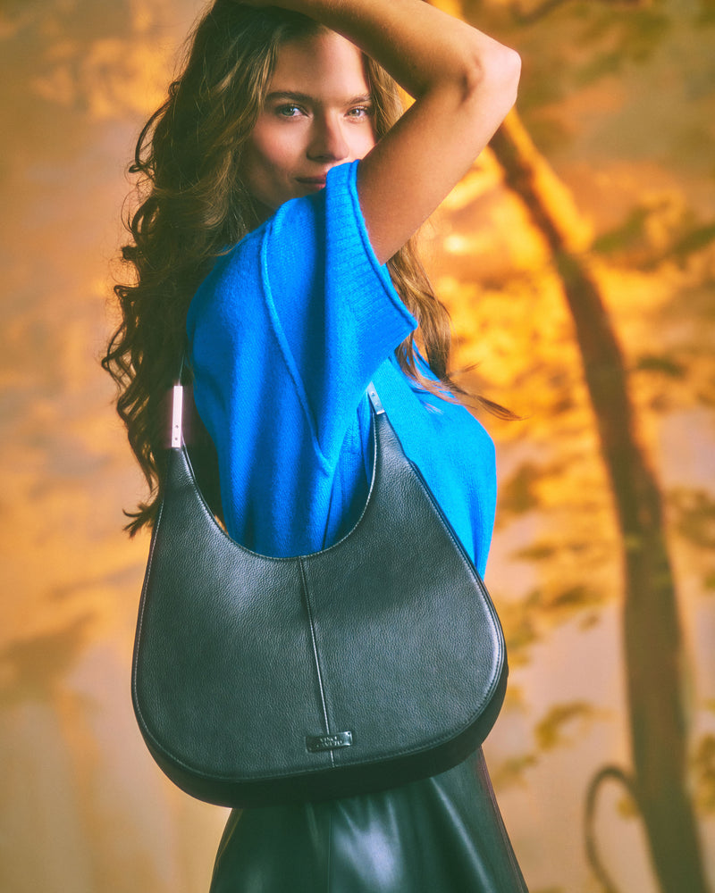 A woman with long hair in a blue top lifts her arm, showcasing the Vince Camuto Noli Hobo Bag— a spacious black purse—over her shoulder. The background features warm, blurred tones and tree silhouettes.