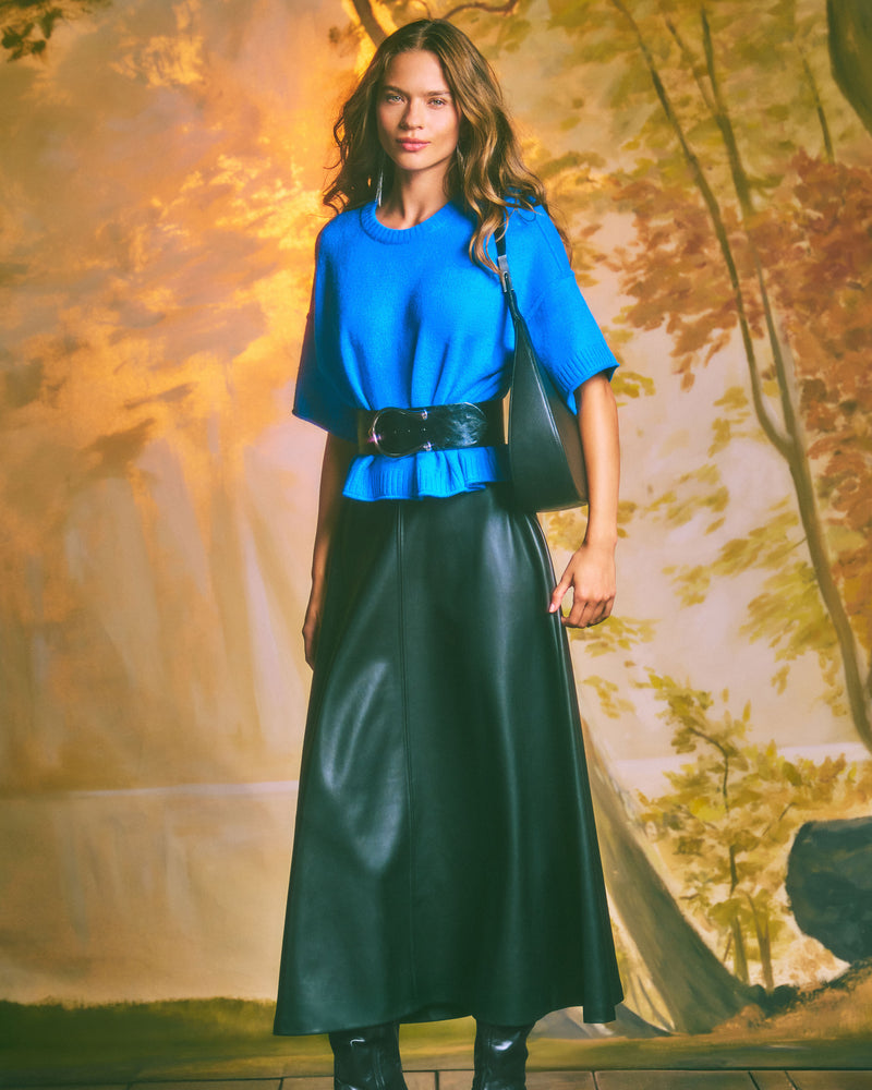 A woman in a bright blue top and a long black leather skirt stands indoors against a backdrop of autumn trees, smiling at the camera. She carries the Vince Camuto Noli Hobo Bag and completes her look with black boots.