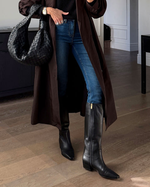 A person wearing a long dark coat, blue jeans, and the Vince Camuto Pavla Knee High Boot—sleek black boots with a block heel—holds a black woven handbag. Only their lower half is visible, standing on a wooden floor in a modern, bright room.