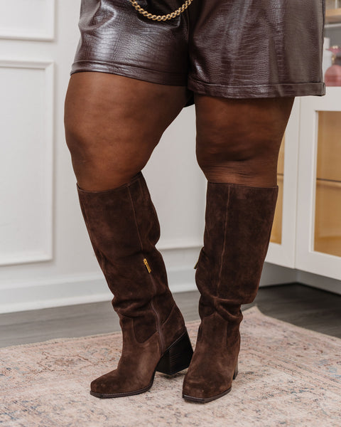 A person wearing the Sangeti Extra Wide Calf Boot by Vince Camuto—brown knee-high suede boots with a stylish block heel—and brown faux leather shorts stands on a light-patterned rug in a modern room.