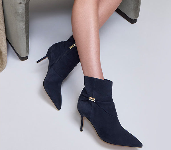 A pair of navy blue suede ankle boots with pointed toes and slim stiletto heels, shown on a model's feet. The boots feature a decorative gold-tone chain detail on the side and a gold zipper on the back.