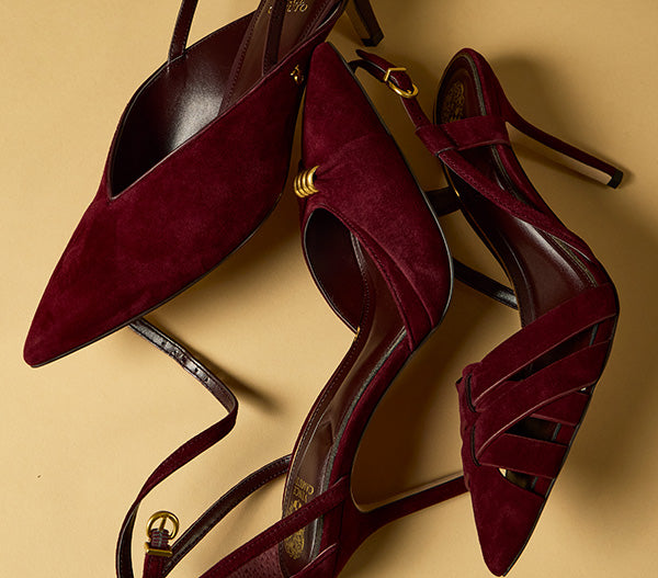 A pair of pointed-toe high heels in deep burgundy suede with gold-tone hardware, arranged on a warm beige background.