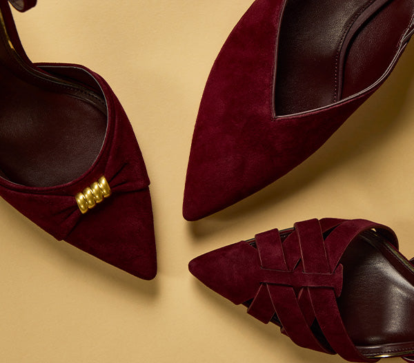 Three pointed-toe suede mule heels in deep burgundy, arranged on a warm beige background. One shoe features a small gold bow with three gold beads, while the others show different strap configurations.