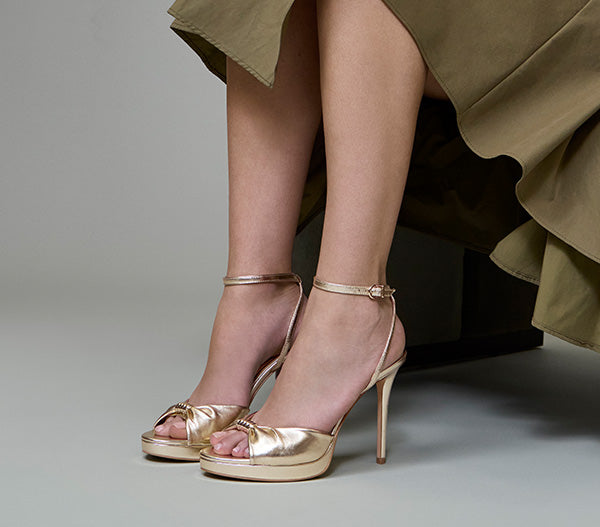 A close-up of a model's feet wearing metallic gold platform high-heel sandals with ankle straps, styled with a draped olive green skirt against a neutral gray studio background.